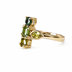A yellow gold ring featuring a cluster of green gemstones, including a marquise-cut lime green stone, a rectangular emerald-cut green stone, and a round dark green stone, set on a polished gold band against a white background.