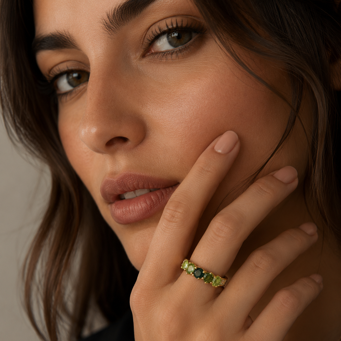 A close-up lifestyle shot of a woman wearing a delicate gold band ring adorned with small green and dark blue gemstones, resting on her ring finger.