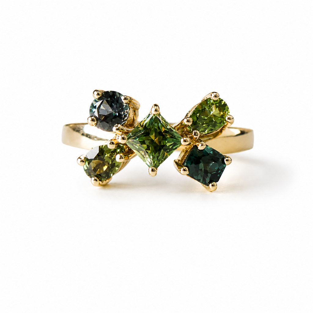 A gold ring featuring a bow-like cluster of green gemstones, including a central square green stone, two marquise green stones, and two oval dark green stones, set on a plain white background.