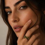 A close-up lifestyle shot of a woman wearing a gold ring with green and blue gemstones on her finger, set against a soft beige background.