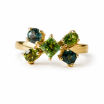 A gold ring featuring a bow-like cluster of green gemstones, including a central square-cut green stone and four surrounding round-cut green and blue-green stones, set against a plain white background.