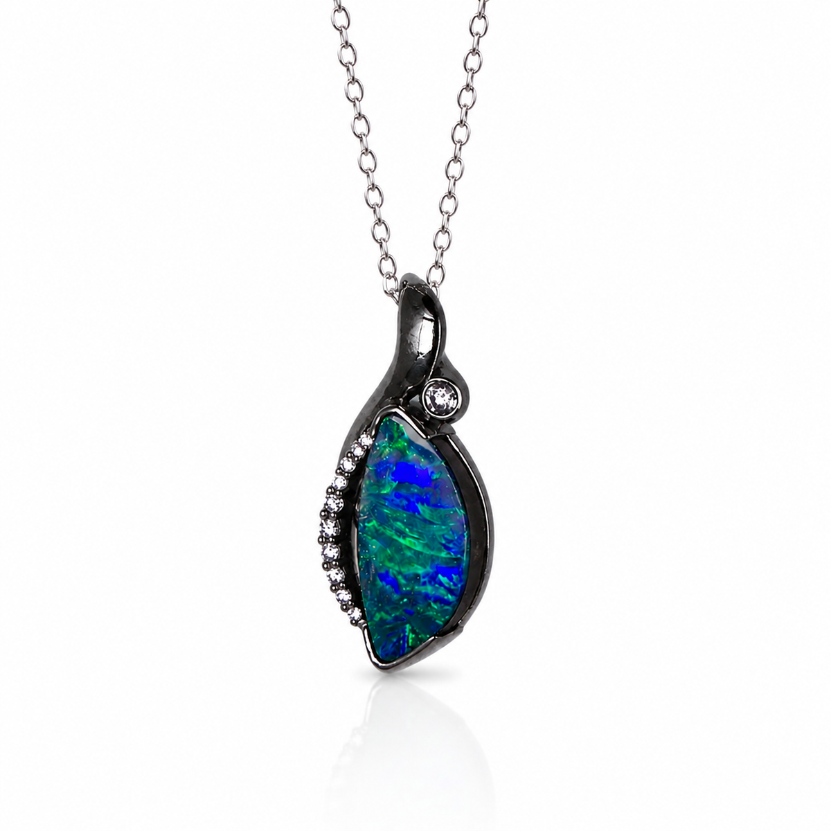 A silver pendant necklace featuring a teardrop-shaped opal stone with vibrant blue and green play-of-color, accented by small clear pavé stones along the left edge and a single round clear stone near the top, suspended from a delicate silver chain on a white background.