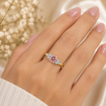 Gold ring with a pink gemstone on a hand with blurred flowers in the background